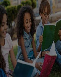 Young school children with backpacks and notebooks sitting outdoors in the grass